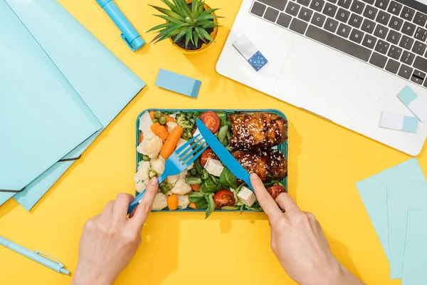 Vue recadrée de la femme tenant fourchette sur la boîte à lunch avec de la nourriture près d'un ordinateur portable et des fournitures de bureau — Photo de stock