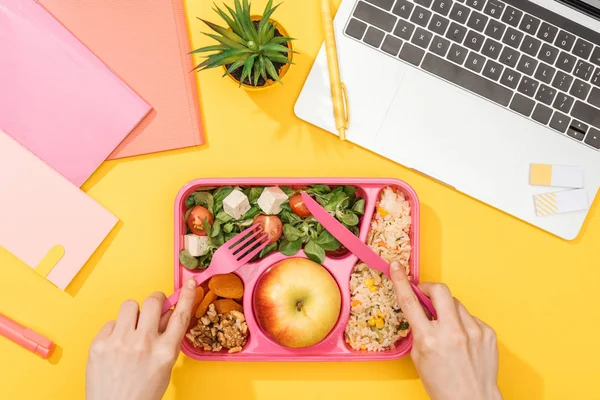 Vista cortada da mulher segurando garfo sobre a lancheira com comida perto de laptop e material de escritório — Fotografia de Stock