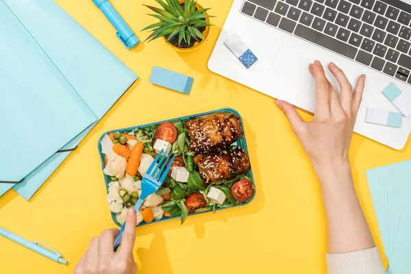 Vista cortada da mulher que trabalha com laptop perto da caixa de almoço e material de escritório — Fotografia de Stock