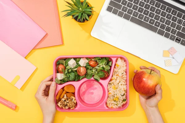 Cropped view of woman holding lunch box with food near laptop and office supplies — Stock Photo