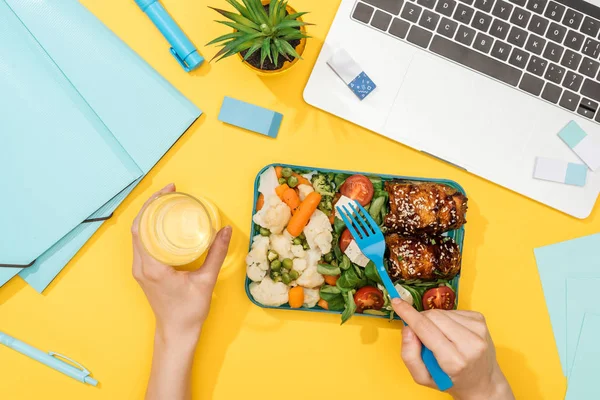 Vista recortada de la mujer sosteniendo lonchera con comida y vaso de agua cerca de la computadora portátil y suministros de oficina - foto de stock