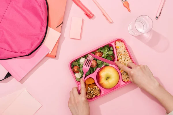 Обрезанный вид женских рук с пластиковой посудой над обеденной коробкой с едой возле рюкзака и канцелярских принадлежностей — стоковое фото