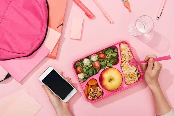 Cropped view of woman with smartphone near lunch box, backpack and stationery — Stock Photo