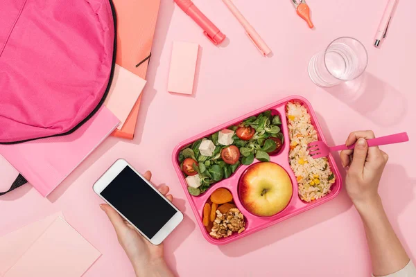 Vista recortada de la mujer con teléfono inteligente cerca de lonchera, mochila y papelería - foto de stock