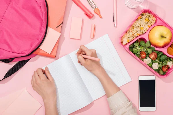 Vista recortada de la mujer escribiendo en el cuaderno cerca de la mochila y artículos de papelería - foto de stock