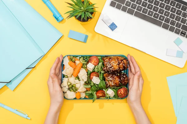 Vue recadrée de la femme tenant boîte à lunch avec de la nourriture près d'un ordinateur portable avec papeterie — Photo de stock