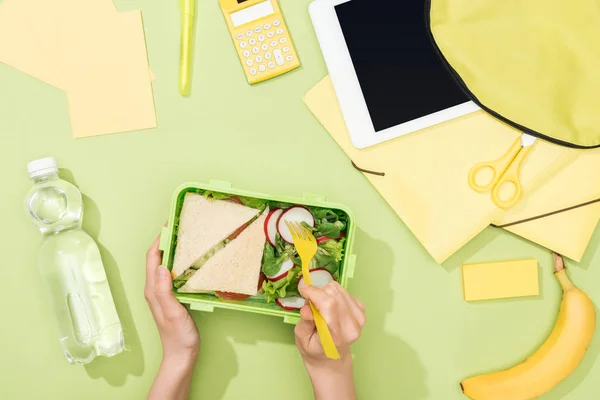 Cropped view of woman hands with plastic utensils over lunch box with food near backpack, digital tablet, bottle of water and stationery — Stock Photo