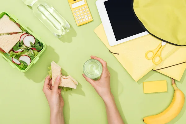 Cropped view of woman hands with sandwich and glass of water near lunch box, backpack, digital tablet, bottle of water and stationery — Stock Photo