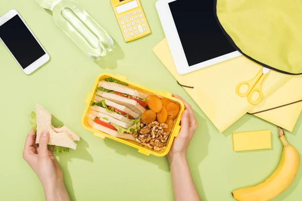 Vista cortada das mãos da mulher com sanduíche, utensílios de plástico ao longo da lancheira com comida perto da mochila, tablet digital, garrafa de água e artigos de papelaria — Fotografia de Stock