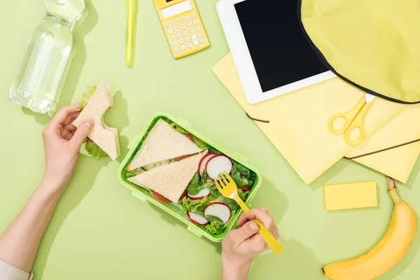 Vista recortada de manos de mujer con sándwich, utensilios de plástico sobre la lonchera con comida cerca de la mochila, tableta digital, botella de agua y papelería - foto de stock