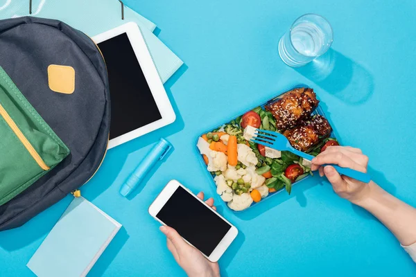 Vista cortada das mãos da mulher com garfo e smartphone perto da lancheira com comida, mochila, tablet digital, vidro de água e papelaria — Fotografia de Stock