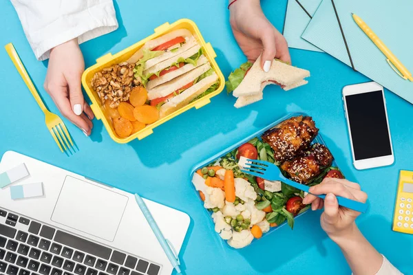 Vue recadrée de deux femmes tenant des fourchettes sur des boîtes à lunch avec de la nourriture près d'un ordinateur portable — Photo de stock