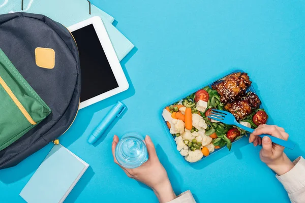 Cropped view of woman holding lunch box and glass of water near backpack with folders, stationery and digital tablet — Stock Photo