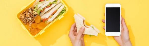 Vista recortada de la mujer sosteniendo sándwich y teléfono inteligente cerca con comida - foto de stock