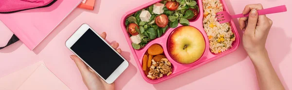 Vista recortada de la mujer sosteniendo teléfono inteligente y tenedor en las manos cerca de la lonchera con comida - foto de stock