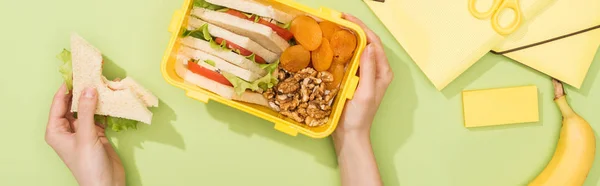 Cropped view of woman holding sandwich, lunch box with food near banana and stationery — Stock Photo