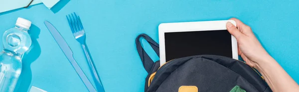 Vista recortada de la mujer embalaje tableta digital en la mochila cerca de tenedor y cuchillo - foto de stock