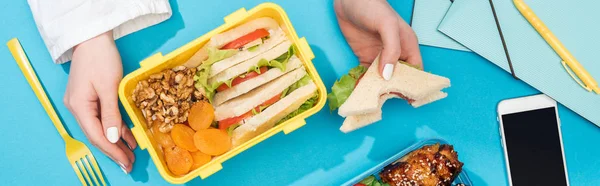 Vue recadrée de la femme avec boîte à lunch et sandwich à la main près de smartphone et fournitures de bureau — Photo de stock