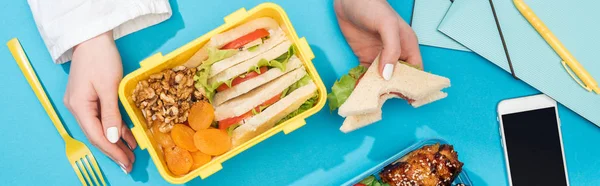 Vista recortada de la mujer con lonchera y sándwich en la mano cerca de teléfonos inteligentes y suministros de oficina - foto de stock