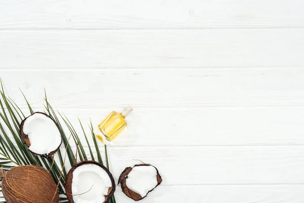 Vista superior de óleo de coco em garrafa na folha de palma verde perto de metades de coco na superfície de madeira branca — Fotografia de Stock