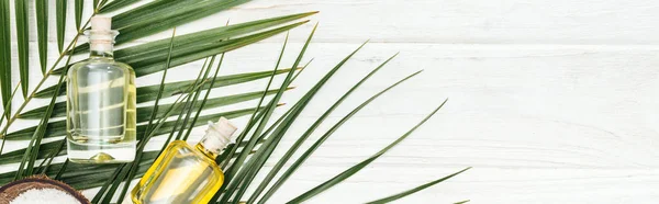 Top view of coconut oil in bottles on green palm leaf on white wooden surface, panoramic shot — Stock Photo