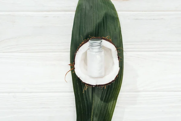 Vista superior do produto de beleza de coco em garrafa na metade de coco e folha de palma verde na superfície de madeira branca — Fotografia de Stock