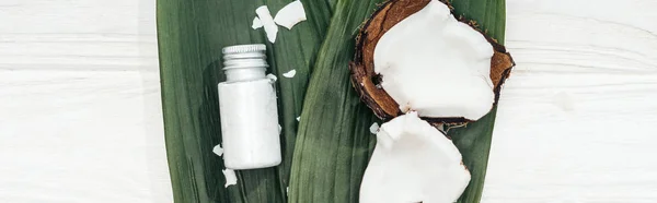 Vista superior del producto de belleza de coco en botella en hojas de palma verde con hojuelas de coco en superficie de madera, plano panorámico - foto de stock