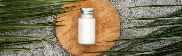 Top view of coconut beauty product in bottle on wooden board near palm leaves on grey textured background, panoramic shot — Stock Photo