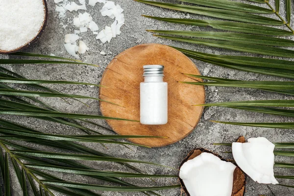 Vista superior do produto de beleza de coco em garrafa na placa de madeira perto de folhas de palma no fundo texturizado cinza — Fotografia de Stock