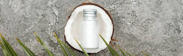 Top view of cream in bottle on coconut half on grey textured background with palm leaf, panoramic shot — Stock Photo