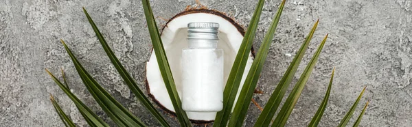 Top view of cream in bottle on coconut half on grey textured background with green palm leaf, panoramic shot — Stock Photo