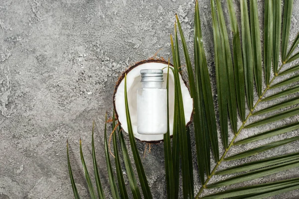 Vue du dessus de la crème en bouteille sur la moitié de noix de coco sur fond texturé gris avec feuille de palme verte — Photo de stock