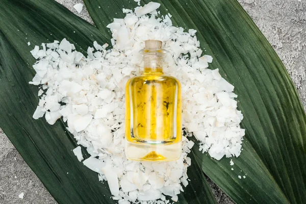 Вид зверху на кокосову олію в пляшці на кокосовій стружці та листі долоні на сірому текстурованому фоні — стокове фото