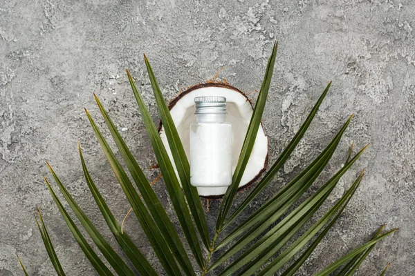 Draufsicht auf Creme in der Flasche auf Kokosnusshälfte auf grauem strukturiertem Hintergrund mit Palmblatt — Stockfoto