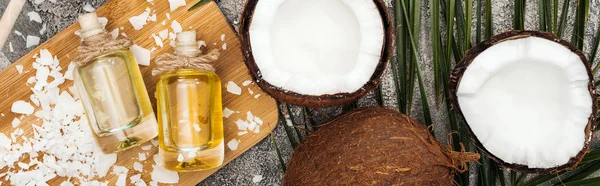 Top view of coconut oil in bottles on wooden board on grey textured background with palm leaf and coconuts, panoramic shot — Stock Photo