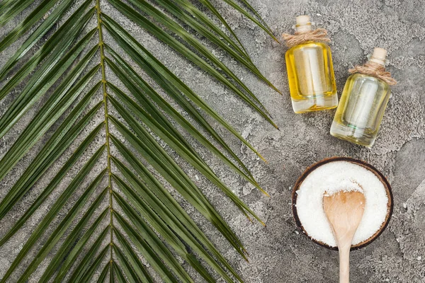 Vue de dessus des copeaux de noix de coco avec cuillère en bois près de l'huile en bouteilles sur fond gris texturé avec feuille de palme — Photo de stock