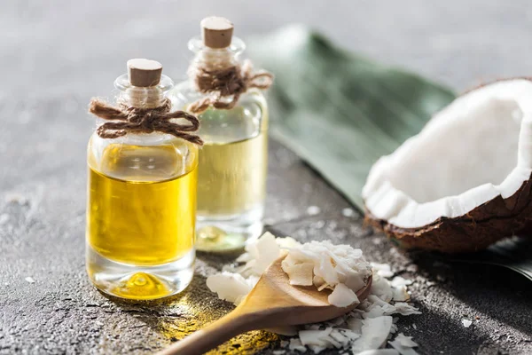 Enfoque selectivo de botellas con aceite de coco cerca de la mitad de coco y virutas en cuchara de madera sobre fondo gris - foto de stock