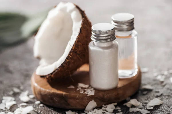 Selective focus of bottles with coconut beauty products on wooden board near coconut half on grey textured background — Stock Photo