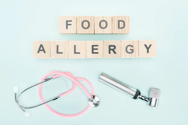 Vista superior do estetoscópio rosa perto de blocos de madeira com letras de alergia alimentar e dermatoscópio no fundo azul — Fotografia de Stock