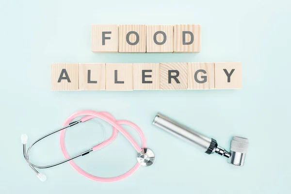 Vista superior del estetoscopio rosa cerca de bloques de madera con letras de alergia alimentaria y dermatoscopio sobre fondo azul - foto de stock