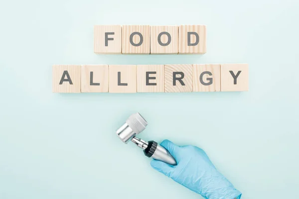 Cropped view of doctor in latex glove holding dermatoscope near wooden blocks with words food allergy on blue — Stock Photo