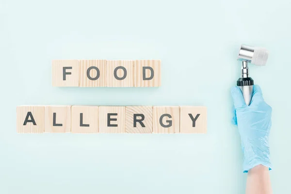 Cropped view of doctor holding dermatoscope near wooden blocks with food allergy inscription on blue — Stock Photo