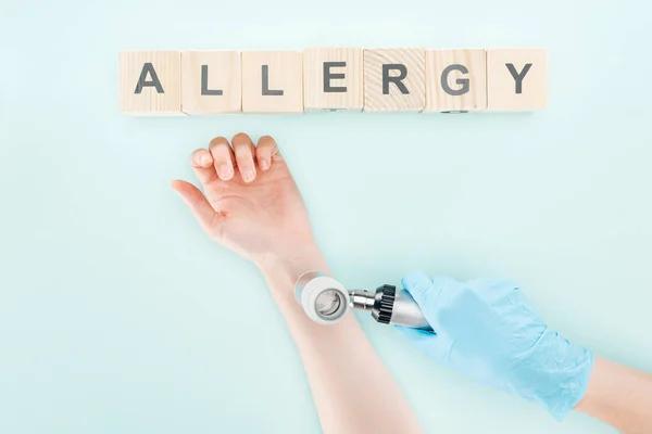 Vista recortada del médico examinando la mano de la mujer con dermatoscopio cerca de bloques de madera con alergia palabra aislada en azul - foto de stock