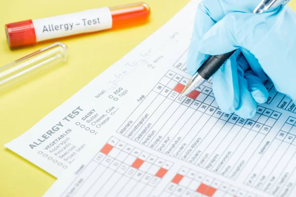 Vista cortada da mulher em luva de látex preenchendo papéis perto de tubos de ensaio com letras de teste de alergia isoladas em amarelo — Fotografia de Stock