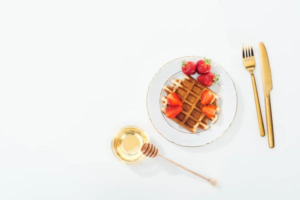 Vista superior de waffle com morangos na placa perto da tigela com mel e mergulhador de madeira, e talheres em branco — Fotografia de Stock