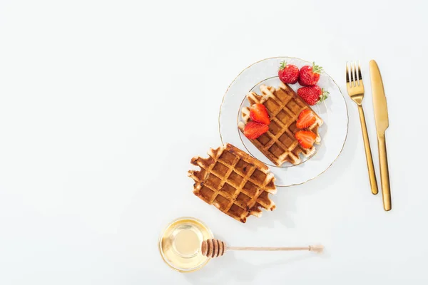 Vista superior de waffles com morangos na placa perto de talheres, tigela com mel e mergulhador de madeira no branco — Fotografia de Stock