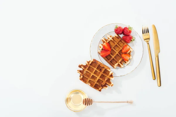 Vista superior de gofres con fresas en el plato cerca de cubiertos, tazón con miel y cazo de madera en blanco - foto de stock