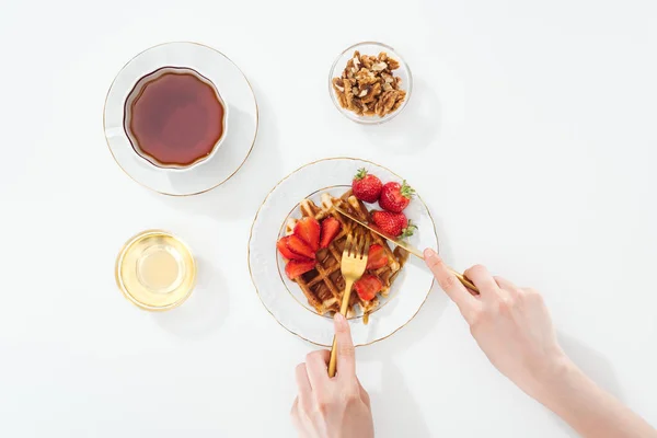 Abgeschnittene Ansicht der Frau Schneiden von Waffel auf Teller in der Nähe Tasse mit Tee und Schüssel auf weiß — Stockfoto