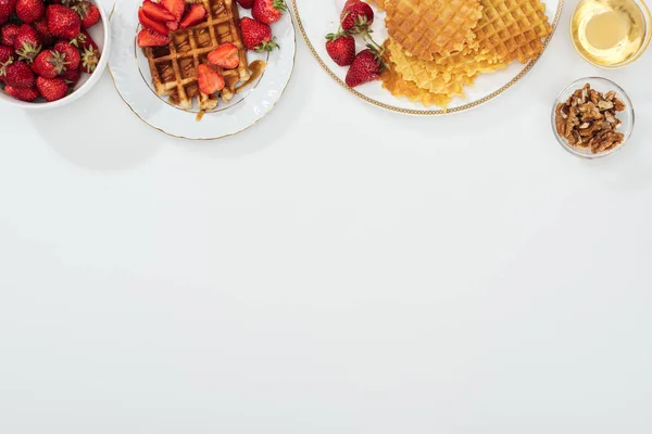 Top view of waffles and strawberries on plated near bowls with honey and nuts on white — Stock Photo