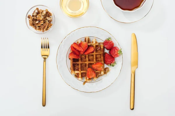 Vista dall'alto della colazione servita con waffle e fragole sul piatto, miele e lendini in ciotole, tazza di tè vicino alla forchetta e coltello su bianco — Foto stock